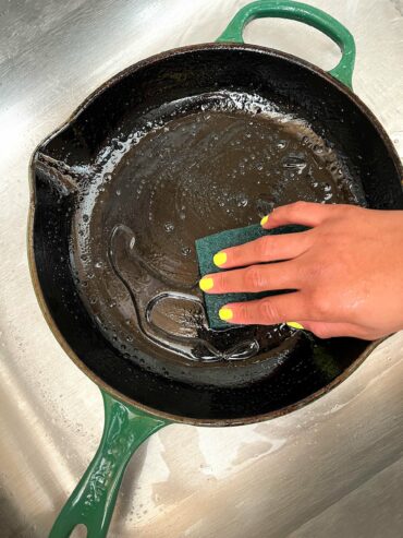 A cast iron skillet being scrubbed in a sink with hot water.